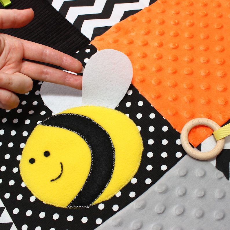 Puede incluir: Una colorida manta sensorial para beb&eacute;s con un aplique de abeja amarilla y negra sonriente. La manta presenta varias texturas y patrones, incluyendo lunares, un dise&ntilde;o en zigzag y un anillo de madera con una cinta amarilla.