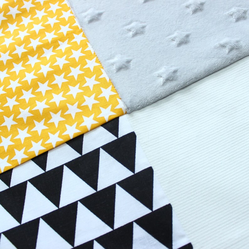 May include: A patchwork of fabrics in yellow with white stars, gray with embossed stars, black and white triangles, and a light blue ribbed fabric. The fabrics are sewn together at angles, creating a geometric design.
