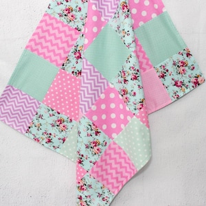 Peut inclure: Un textile patchwork avec des carrés de tissus roses, bleu clair et lavande. Le motif comprend des motifs floraux, des pois, des chevrons et des imprimés étoiles. Le textile est probablement une courtepointe ou une couverture.