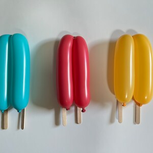 May include: Three balloon popsicles on wooden sticks. The popsicles are blue, red, and yellow.