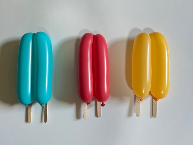 May include: Three balloon popsicles on wooden sticks. The popsicles are blue, red, and yellow.