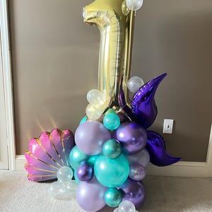 May include: A gold number one balloon with purple and teal balloons, a purple mermaid tail, and a pink seashell balloon. The balloons are arranged in a cluster.