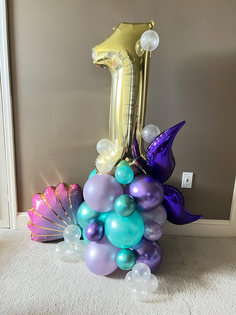 May include: A gold number one balloon with purple and teal balloons, a purple mermaid tail, and a pink seashell balloon. The balloons are arranged in a cluster.