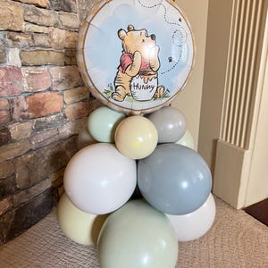 Puede incluir: Un arreglo de globos con un globo redondo de aluminio de Winnie the Pooh encima de un grupo de globos de colores pastel. El globo de aluminio tiene un fondo azul claro y la palabra "Hunny" en el bote de miel.