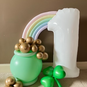 May include: A green pot of gold balloon with gold balloons spilling out, a rainbow balloon arch, a white number one balloon, and a green shamrock balloon.