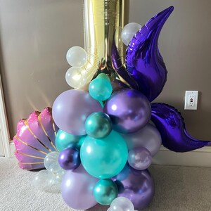 May include: A gold number one balloon with purple, teal, and lavender balloons, a purple tail fin, and a pink seashell balloon.