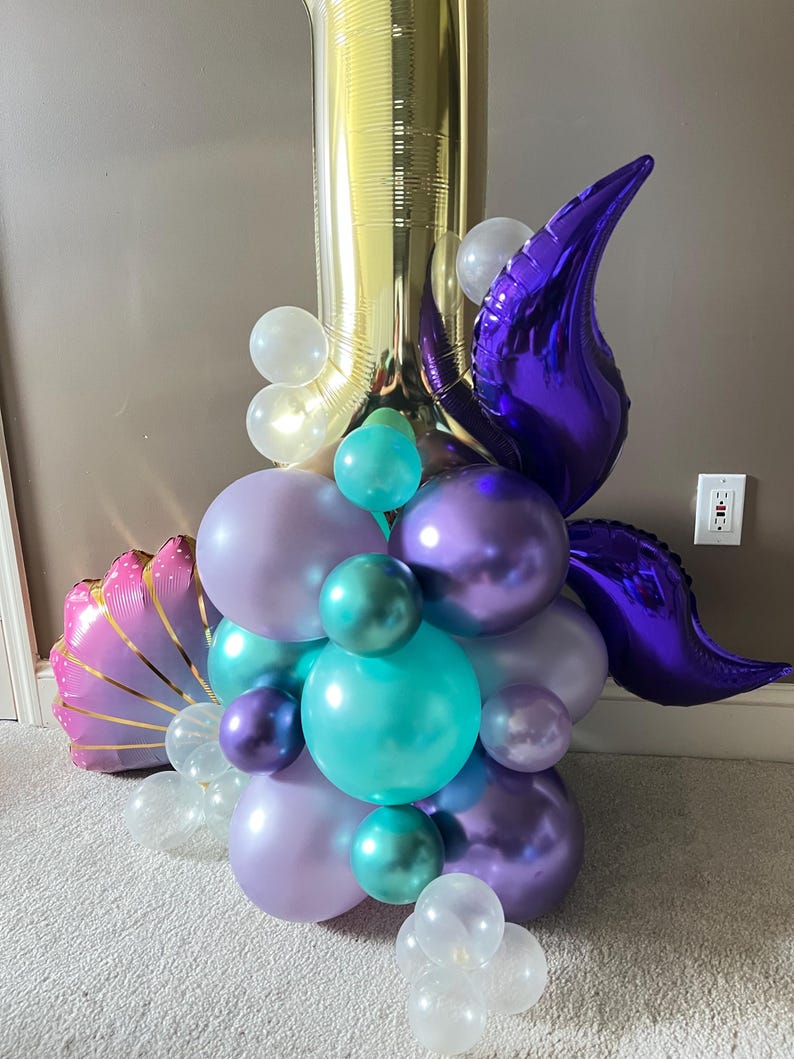 May include: A gold number one balloon with purple, teal, and lavender balloons, a purple tail fin, and a pink seashell balloon.