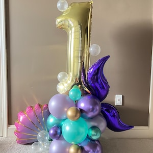 May include: A gold number one balloon with purple and teal balloons, clear balloons, and a pink and gold seashell balloon. The balloons are arranged in a cluster.
