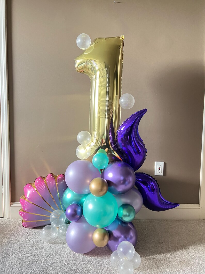 May include: A gold number one balloon with purple and teal balloons, clear balloons, and a pink and gold seashell balloon. The balloons are arranged in a cluster.