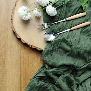 May include: A green fabric napkin with a wooden round serving tray and white flowers. A silver spoon and fork with wooden handles are on top of the napkin.