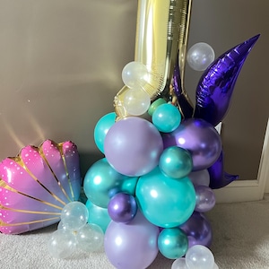 May include: A gold number one balloon with a purple tail and a cluster of purple, teal, and clear balloons. A pink and gold seashell balloon is in the foreground.