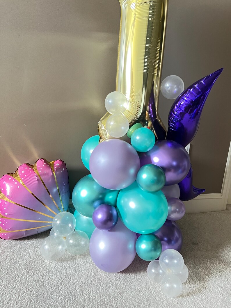 May include: A gold number one balloon with a purple tail and a cluster of purple, teal, and clear balloons. A pink and gold seashell balloon is in the foreground.