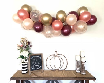 May include: A garland of balloons in shades of burgundy, rose gold, and champagne with gold confetti balloons. The garland is hanging on a white wall. A sign with the text "Fall in Love" is on a table with a wire pumpkin and candles.