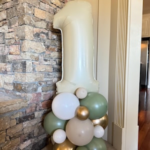 Puede incluir: Un gran globo número uno de color verde pálido se encuentra sobre un grupo de globos blancos, dorados y verdes. Los globos están dispuestos en una exhibición decorativa, adecuada para una celebración de primer cumpleaños o un evento importante.