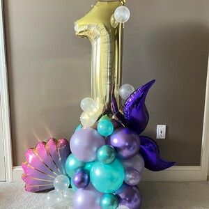 May include: A gold number one balloon with purple and teal balloons, a purple mermaid tail, and a pink seashell balloon. The balloons are arranged in a cluster.