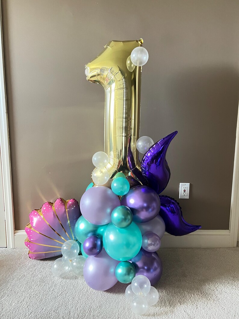May include: A gold number one balloon with purple and teal balloons, a purple mermaid tail, and a pink seashell balloon. The balloons are arranged in a cluster.