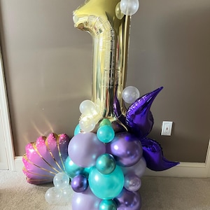 May include: A gold number one balloon with purple, teal, and white balloons arranged around it. The balloons are in the shape of a seashell and a mermaid tail.