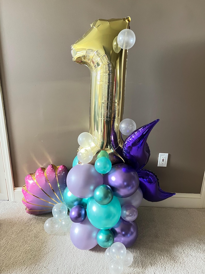 May include: A gold number one balloon with purple, teal, and white balloons arranged around it. The balloons are in the shape of a seashell and a mermaid tail.