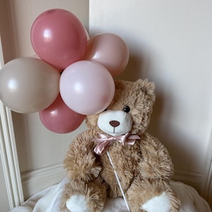 May include: A plush brown teddy bear with a pink bow tie, holding a cluster of five balloons in shades of pink. The balloons are attached to a white stick. The bear is sitting on a white blanket.