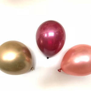 Puede incluir: Tres globos metálicos en dorado, oro rosa y burdeos. Los globos son brillantes y tienen un acabado perlado.