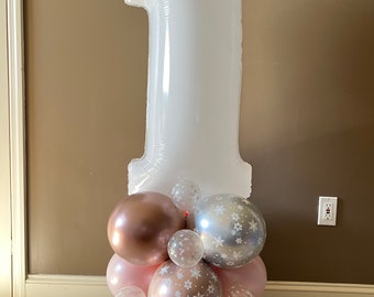May include: A white number one balloon with a cluster of balloons in pink, silver, and rose gold with snowflake patterns. The balloons are arranged in a circular shape around the base of the number one balloon.