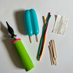May include: A green balloon pump with a black nozzle, two light blue balloons, five colorful rubber bands, a sheet of white adhesive dots, and a pile of wooden craft sticks.