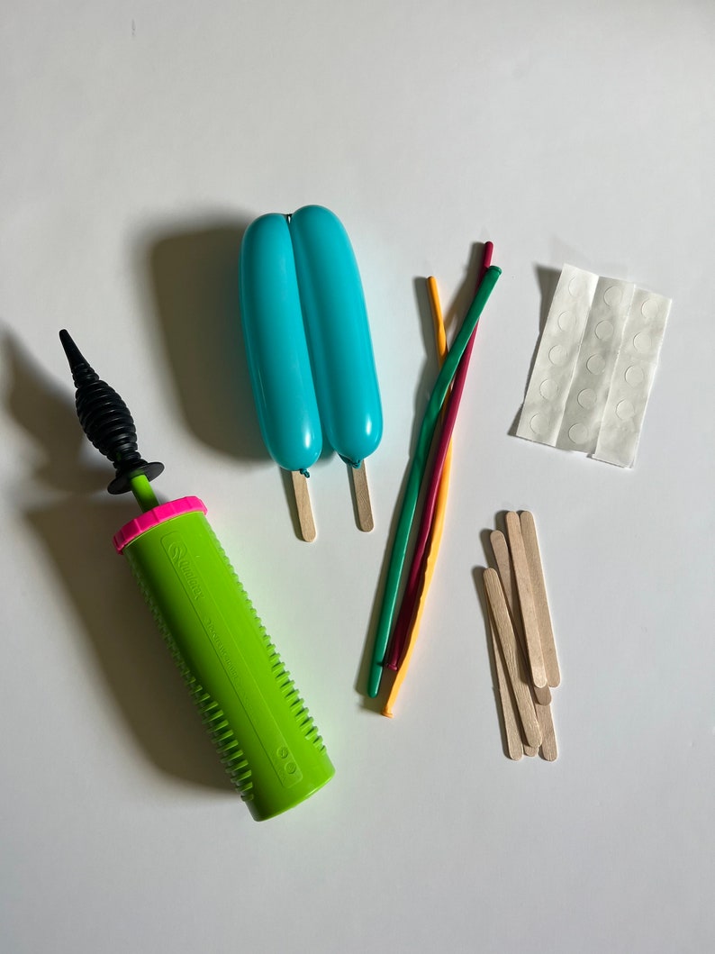 May include: A green balloon pump with a black nozzle, two light blue balloons, five colorful rubber bands, a sheet of white adhesive dots, and a pile of wooden craft sticks.