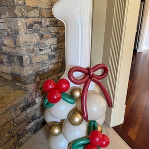 May include: A white number one balloon, part of a balloon arrangement. The base is made of white, gold, and red balloons, with green accents. A large burgundy bow is attached to the top of the balloon arrangement. The arrangement is for a first birthday.