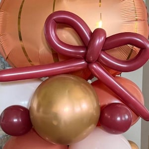 May include: A festive balloon arrangement featuring a large burgundy bow, a gold sphere, and various other balloons in white, burgundy, and peach. A large orange balloon is in the background.