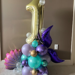 May include: A gold number one balloon with purple and teal balloons, clear balloons, and a purple shell-shaped balloon. The balloons are arranged in a cluster.