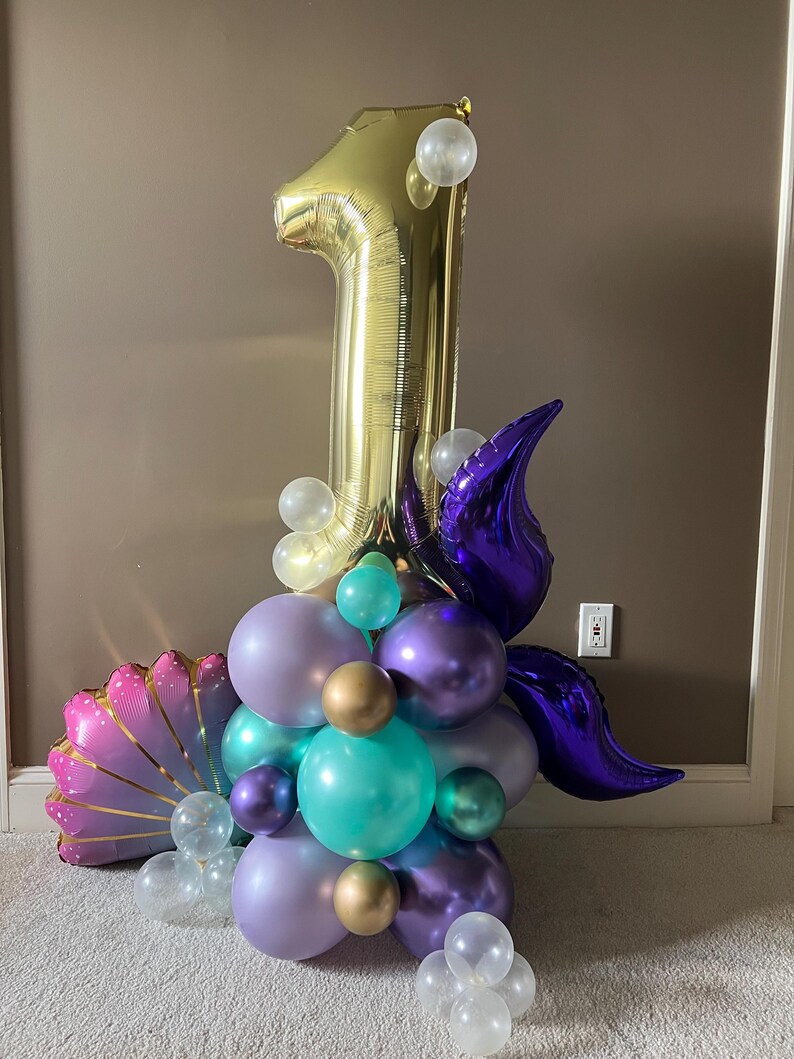 May include: A gold number one balloon with purple and teal balloons, clear balloons, and a purple shell-shaped balloon. The balloons are arranged in a cluster.