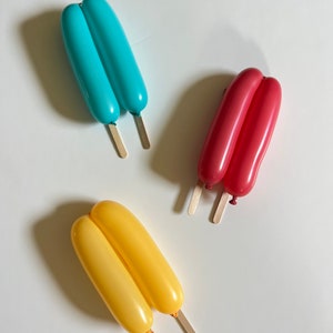 May include: Three balloon animal popsicles on a white background. The popsicles are made of two balloons each, tied together at the top. The popsicles are blue, red, and yellow.