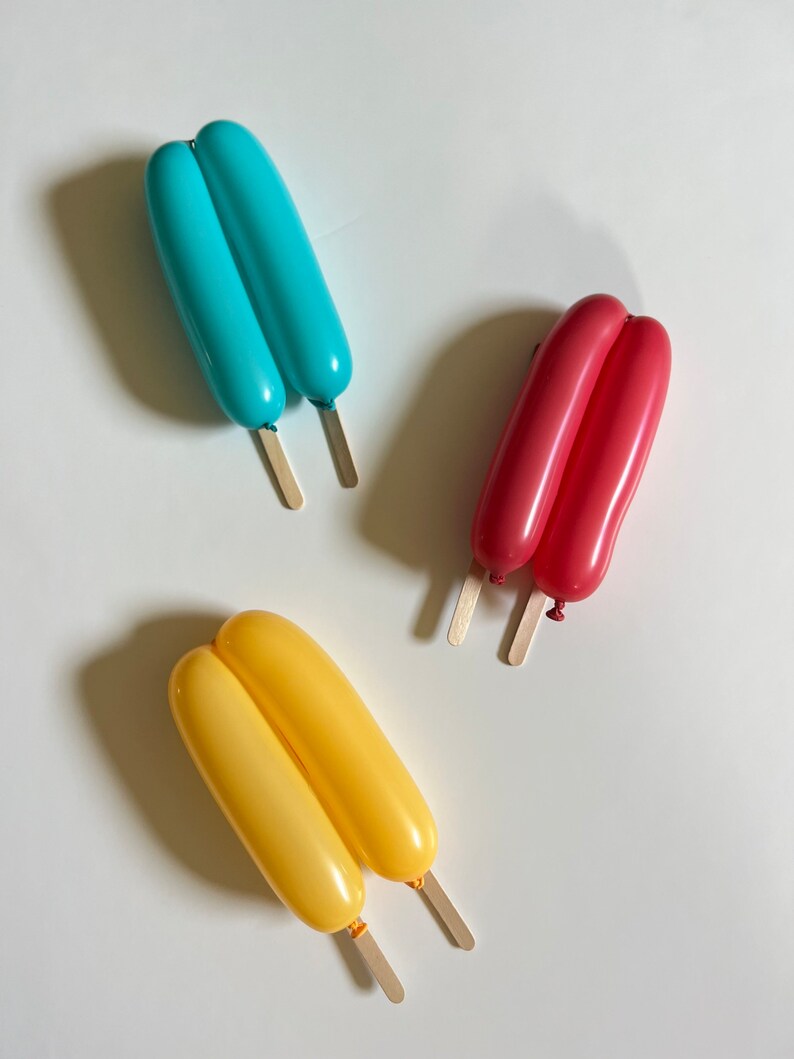 May include: Three balloon animal popsicles on a white background. The popsicles are made of two balloons each, tied together at the top. The popsicles are blue, red, and yellow.