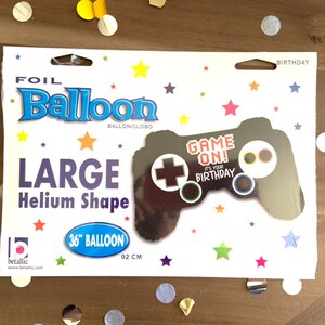 Op de afbeelding: Een grote folieballon in de vorm van een gamecontroller. De zwarte ballon heeft de tekst "GAME ON! IT'S YOUR BIRTHDAY." Het meet 92 cm (36 inch).