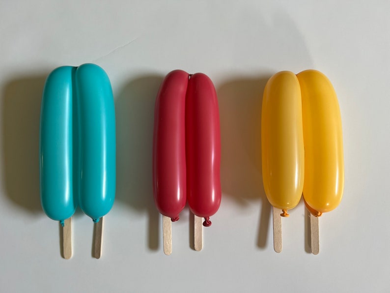 May include: Three balloon popsicles on wooden sticks. The popsicles are blue, red, and yellow.