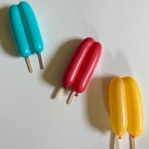 May include: Three balloon popsicles on sticks. The popsicles are blue, red, and yellow.