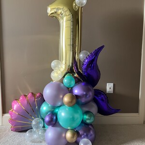 May include: A gold number one balloon with purple, teal, and iridescent balloons arranged around it. The balloons are in the shape of a seashell and a mermaid tail.