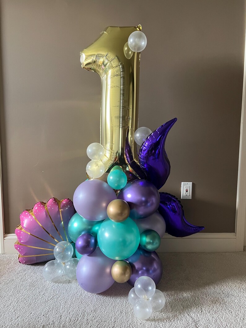 May include: A gold number one balloon with purple, teal, and iridescent balloons arranged around it. The balloons are in the shape of a seashell and a mermaid tail.