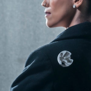 May include: A black blazer with a silver pin featuring a 3D design of a building with a staircase.