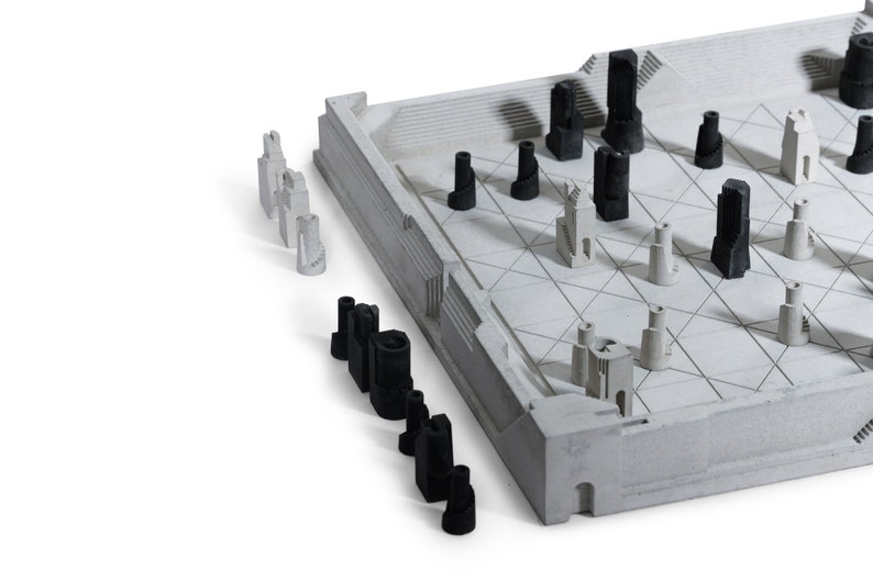 May include: A modern chess set with a concrete-like board and architectural-style pieces. The pieces are in black and white, resembling miniature buildings. The board has a geometric design, and the set is on a white background.