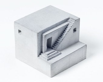 Puede incluir: Una pequeña escultura de hormigón gris de una casa con una escalera que conduce a una puerta. La escultura tiene forma de cubo y presenta un diseño simple y moderno.