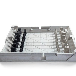 May include: A modern chess set with a concrete-like appearance. The board is a square with a grid pattern, and the pieces are stylized in black and light gray. The set has a minimalist, architectural design.