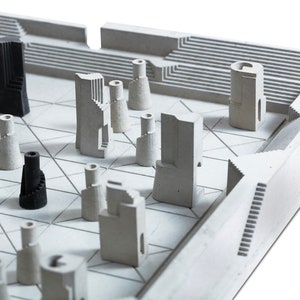 May include: A modern chess set with a geometric design. The board and pieces are primarily light gray, with some pieces in black. The pieces have architectural shapes, resembling miniature buildings and towers. The board has a grid pattern.