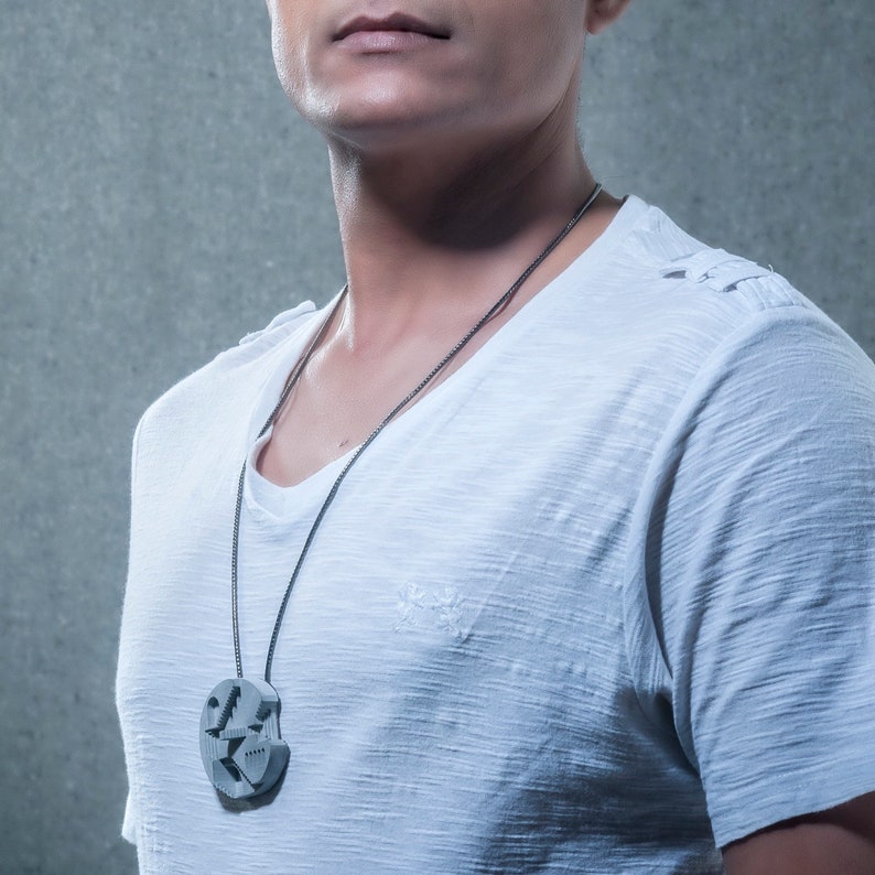 May include: A man wearing a white t-shirt with a silver necklace and a circular pendant. The pendant has a geometric design.