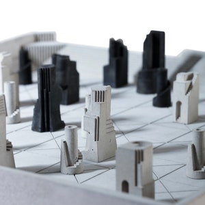 May include: A modern chess set featuring architectural-style pieces in shades of gray and black. The board is a light gray with a geometric pattern. The pieces resemble miniature buildings and towers.