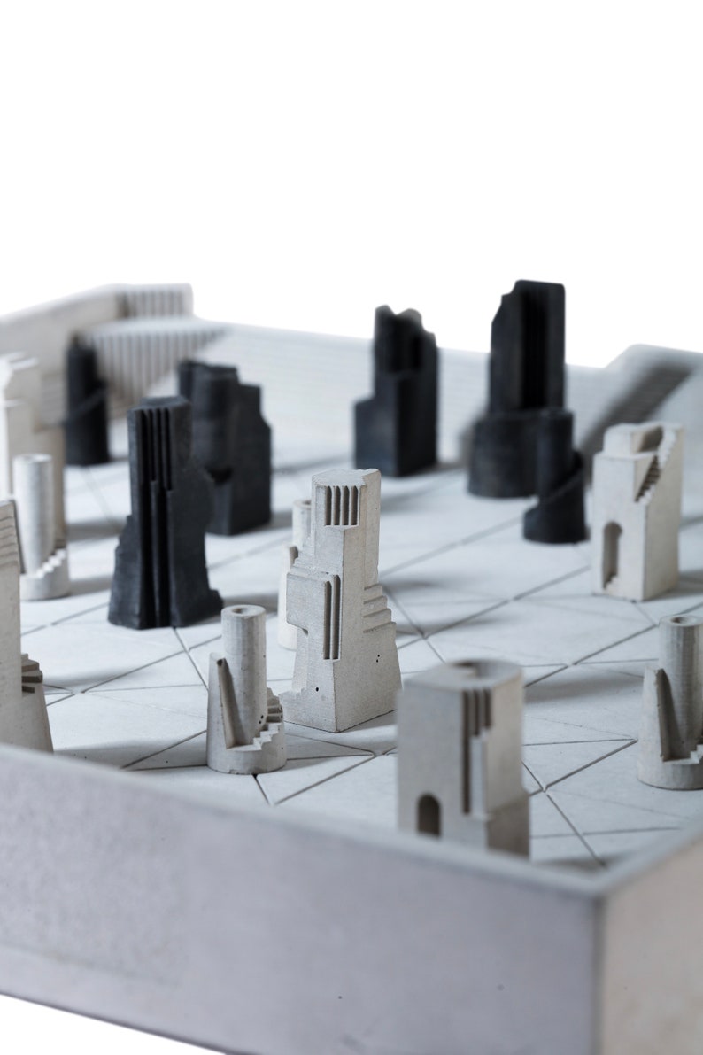 May include: A modern chess set featuring architectural-style pieces in shades of gray and black. The board is a light gray with a geometric pattern. The pieces resemble miniature buildings and towers.