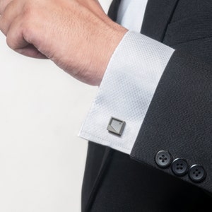 Könnte beinhalten: Ein Mann trägt eine schwarze Sakkojacke mit einem weißen Hemd und Manschettenknöpfen. Die Manschettenknöpfe sind silberfarben und haben ein geometrisches Design.
