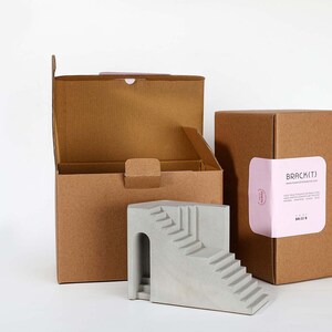 Puede incluir: Una escultura de hormig&oacute;n gris de una escalera con un arco. La escultura est&aacute; empaquetada en dos cajas de cart&oacute;n marr&oacute;n. Una caja tiene una etiqueta rosa con el texto "BRACKETJ" y una direcci&oacute;n de sitio web.