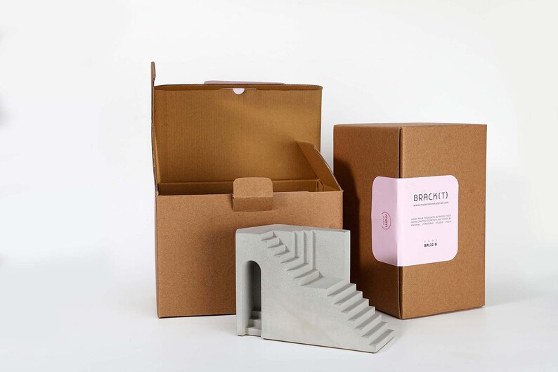Puede incluir: Una escultura de hormig&oacute;n gris de una escalera con un arco. La escultura est&aacute; empaquetada en dos cajas de cart&oacute;n marr&oacute;n. Una caja tiene una etiqueta rosa con el texto "BRACKETJ" y una direcci&oacute;n de sitio web.