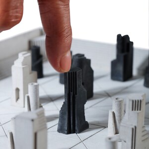 May include: A modern chess set featuring architectural-style pieces in black and white. The pieces are arranged on a white board with a grid pattern. A finger is touching a black piece, highlighting the game's strategic nature.